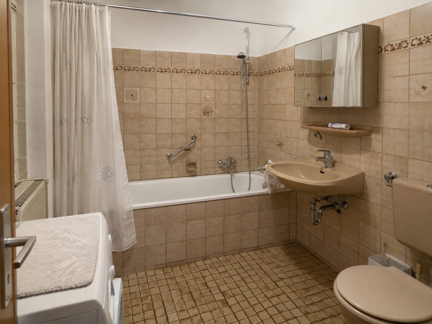 Bad vor der Sanierung: beige Wandfliesen mit Blumenbordüre, Badewanne mit Duschvorhang.